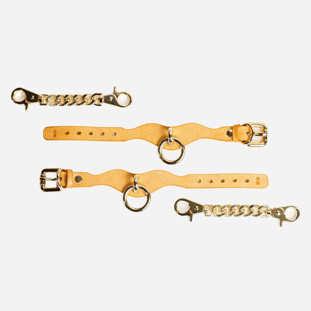 Two wrist cuffs of bright yellow leather opened full length on a white background. Next to them are two chains with crab-claw fasteners.