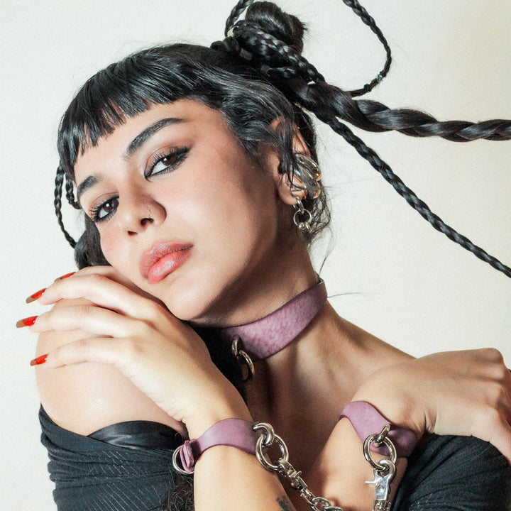 A woman with short black bangs and long braids looks at the camera with her arms crossed. Her wrists have lavender leather BDSM cuffs on them. 