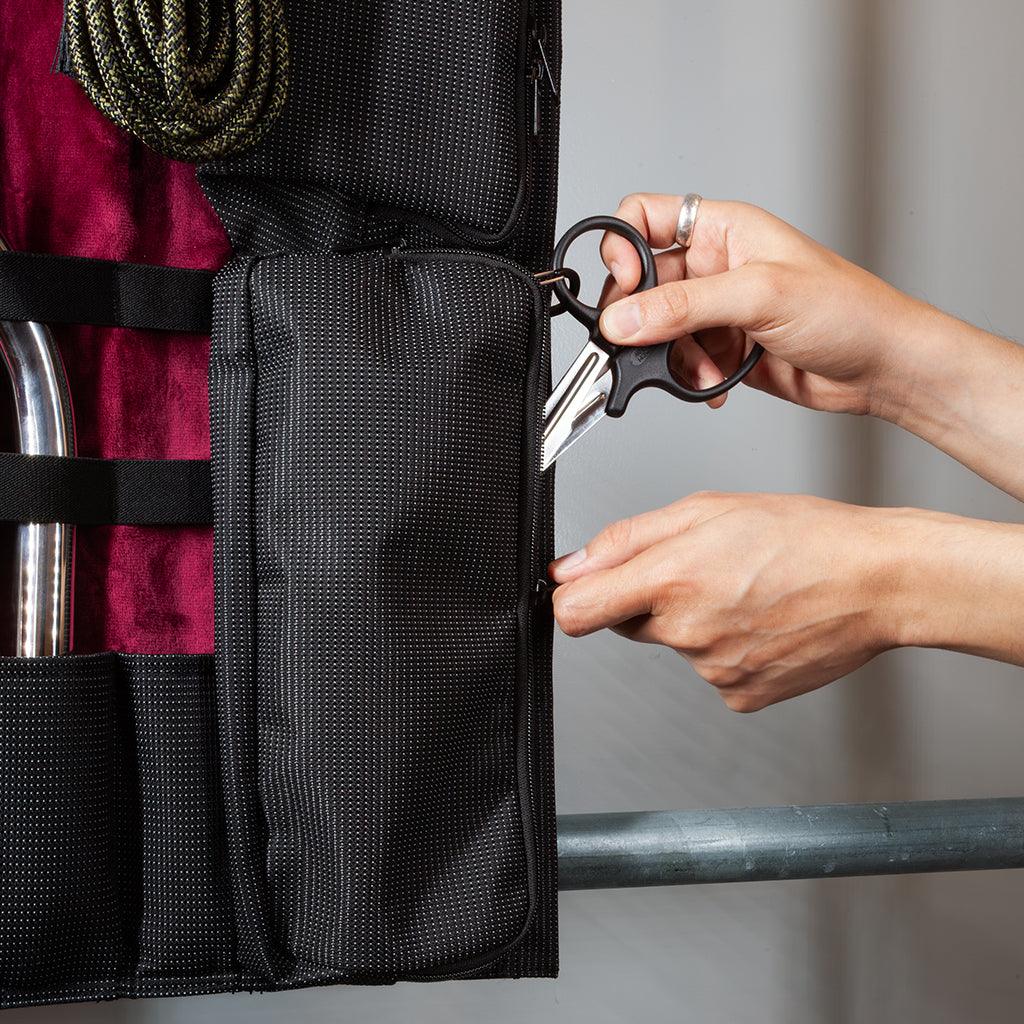 Closeup of one of the pockets on the KinkBag. A person's hands are putting a pair of safety shears into the bag. 