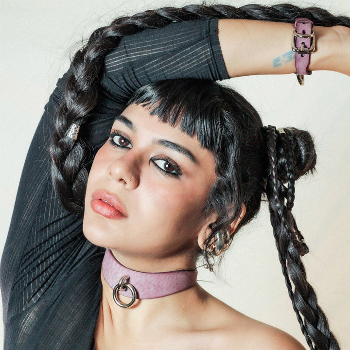 A young woman with short black bangs and long braids looks at the camera, her right arm resting on top of her head. Around her neck is the Neon Coyotes Luxury Leather Day Collar, in hazy pink with a nickel-coated ring in front. 