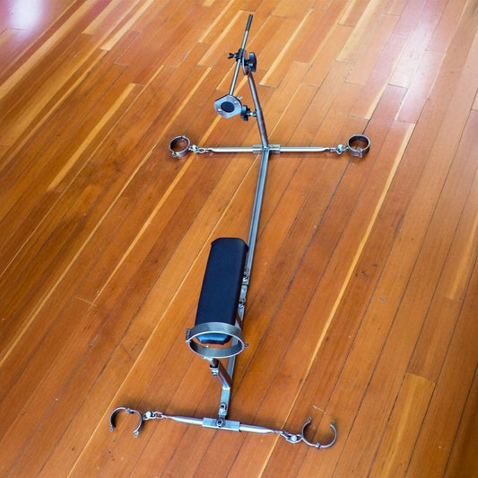 The Stockroom Stockade is shown on a wooden floor. It has 1 vertical bar with 2 horizontal spreader bars with metal cuffs. It has an upright bar with a metal collar and a fucking rod at the other end. The leather stomach support is not upright.