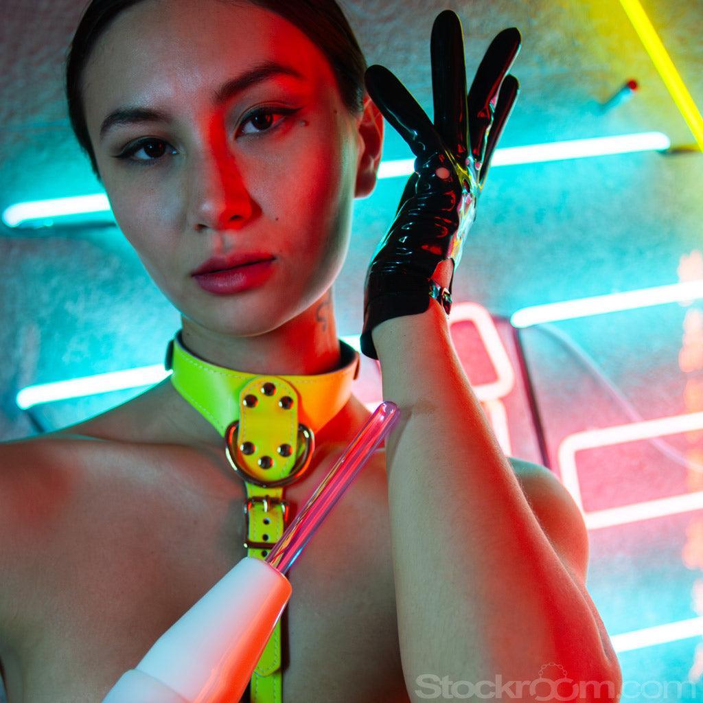A topless woman with her dark hair pulled back, wearing a neon yellow bust harness and black latex driving gloves, is shown from the bust up in front of a wall the neon lights. She holds a neon wand with the KinkLab Probe Neon Wand Electroplay Attachment.