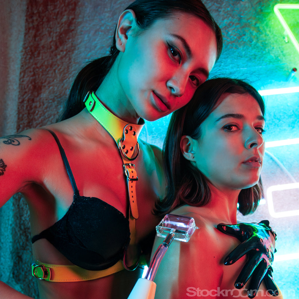 2 women are shown embracing each other in a room lit with neon lights. The first woman is pressing the white handle Neon Wand with roller electrode attachment against the second womans shoulder.