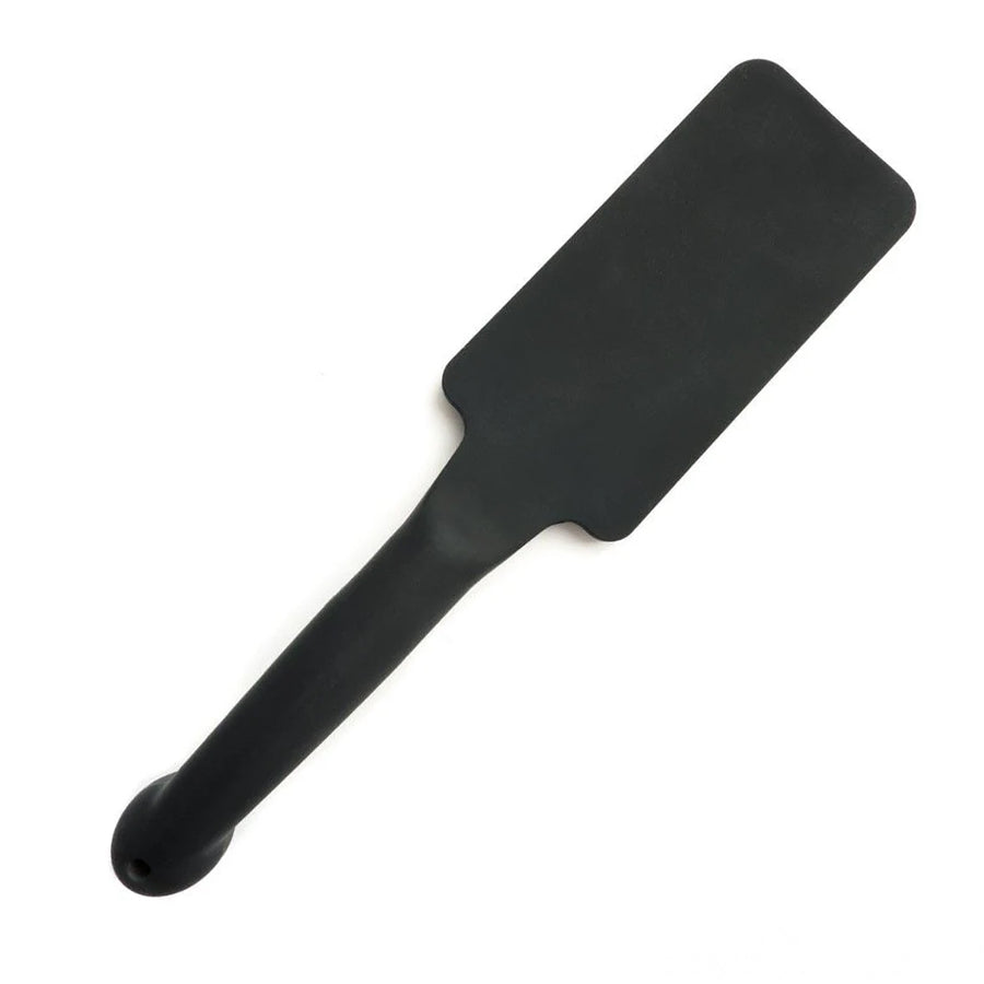 The Tantus Plunge Paddle is displayed against a blank background. The paddle is rectangle-shaped and has a cylindrical handle. It is made of matte black silicone.