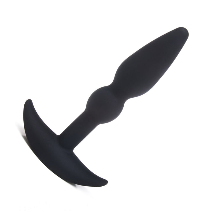 The Tantus Perfect Butt Plug, made of black silicone, is displayed against a blank background. It is thin and tapered and has a small bump at the base before the thin neck of the plug begins. It has a wide, flared base.