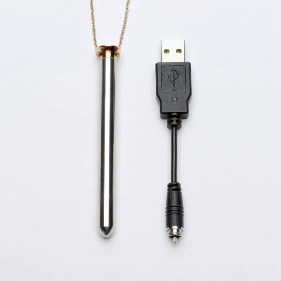The Vesper Pendant Necklace Vibrator in Rose Gold is displayed next to its charger against a blank background. It is a USB charger with a pin plug.