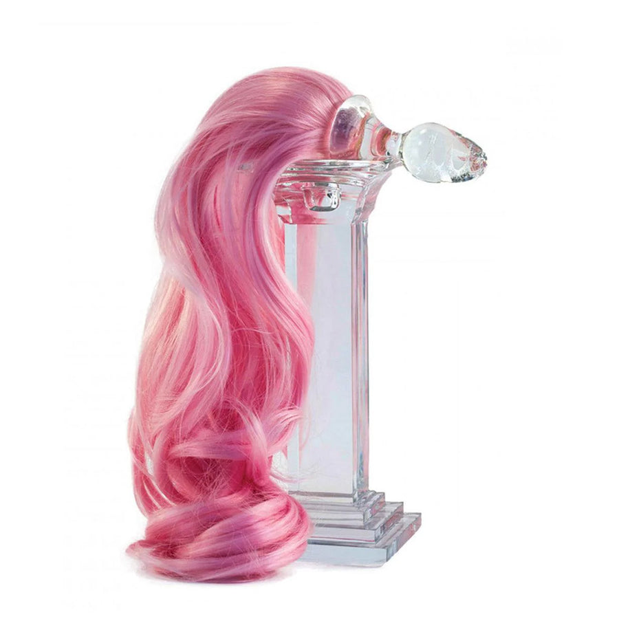 The Pink Detachable Ponytail Glass Butt Plug rests ontop of a clear pedestal in front of a blank background. The tail is draped down the side of the pedestal.