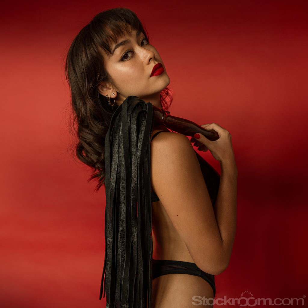 A profile shot of a woman is shown standing in front of a red background. She is holding the Sissoo Rosewood Long Handle Leather Flogger over her shoulder. 