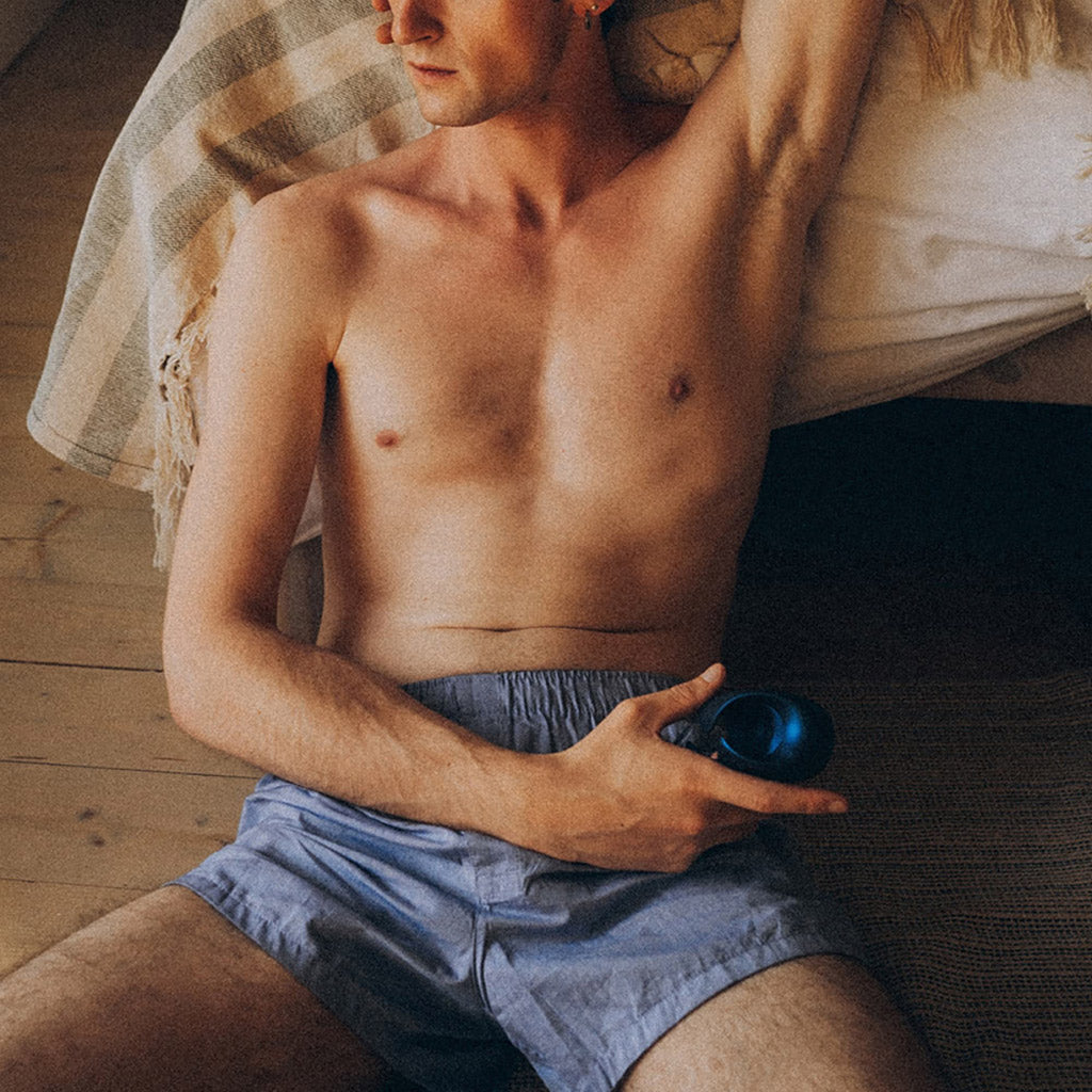 A man reclining on a pillow, shirtless and wearing boxers. In one hand he has the Sub One vibrator. 