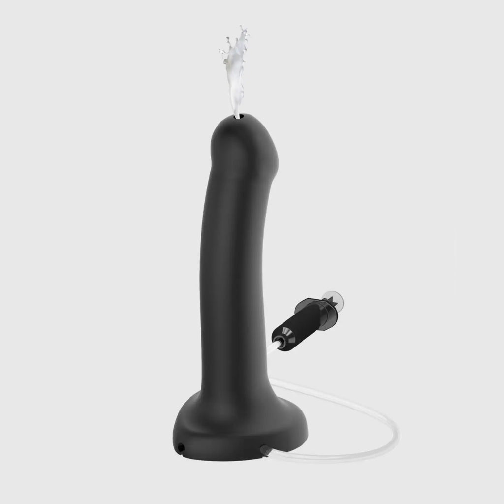 The black strap-on-me semi-realistic squirting dildo is shown on a white background with lube squirting out of the tip. A black syringe and tube are connected to the dildo at the base. 