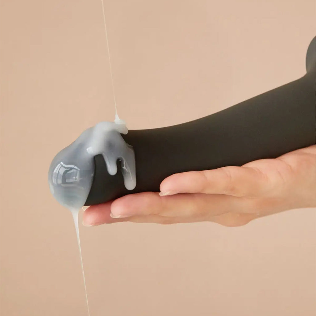 A close-up of a models hand is shown holding the black strap-on-me semi-realistic squirting dildo as its being covered in lube. 