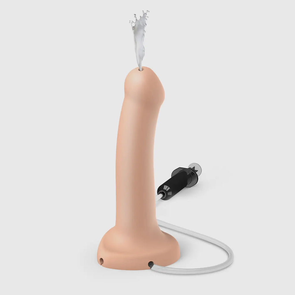 The vanilla colored strap-on-me semi-realistic squirting dildo is shown on a white background with lube squirting out of the tip. A black syringe and tube are connected to the dildo at the base. 