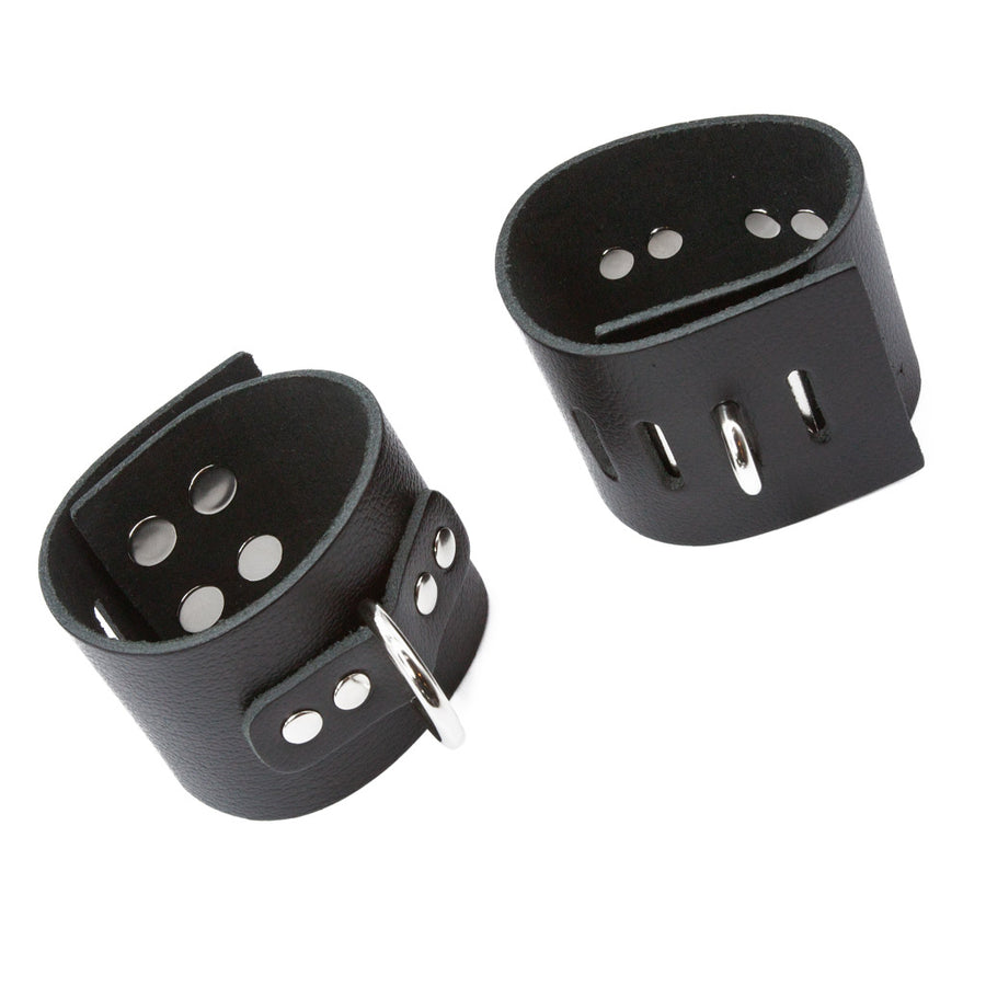 A pair of black leather Bondage Wrist Cuffs with a D-Ring are displayed against a blank background. The cuffs have multiple verticle slots on them for the D-ring closure to fit through.