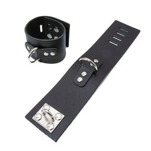 A pair of Bondage Ankle Cuffs w/ D-Ring is displayed against a blank background. The cuffs are made of black leather with a metal D-ring in the center. There are notches on the cuffs and a hasp closure.
