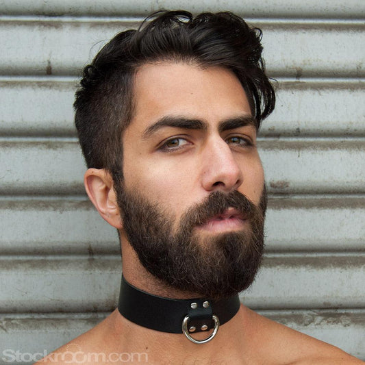 A close-up of a dark-haired man's face is shown in front of a metal wall. He has facial hair and is wearing the Locking Leather Collar. The collar is a wide piece of black leather with a silver D-ring in the front.