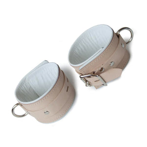 A pair of Institutional Leather BDSM Wrist Restraints are shown against a blank background. The cuffs are made of a wide piece of tan leather with a white leather-lined interior. They each have a silver D-ring and a lockable buckle.