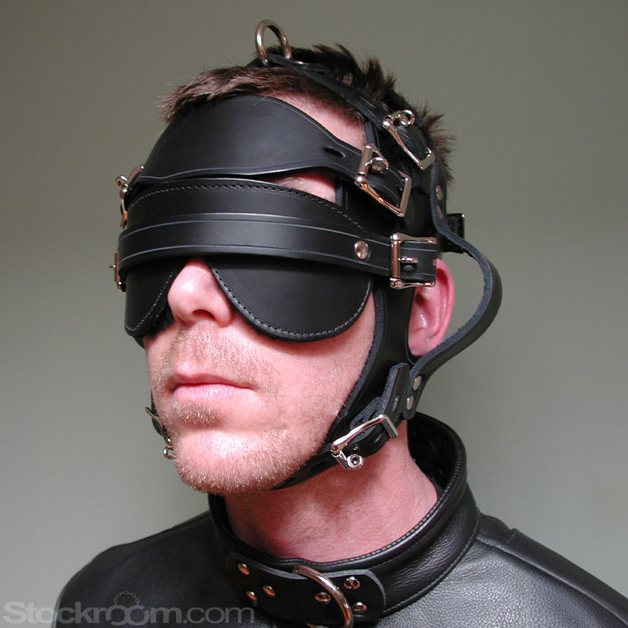 A close-up of a man's head in the Inescapable Head Harness is shown in front of a grey background. The black leather harness goes around the sides of his head and has loops near the ears. It also has an attached blindfold.
