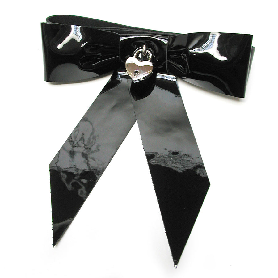 The black Patent Leather Bow Wrist Restraint is shown from the front against a blank background. It is a large bow made of shiny black patent leather. There is a silver heart padlock in the center of the bow.