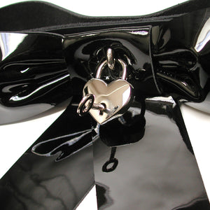 A close-up of the center of the black Patent Leather Bow Wrist Restraint is shown against a blank background. A key is shown inserted into the keyhole of the heart padlock.