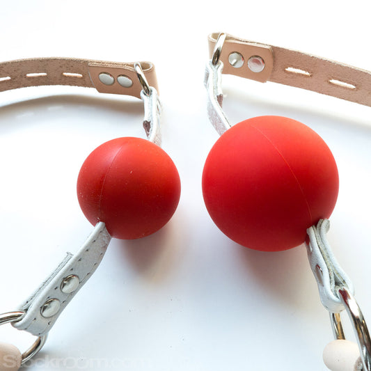 Two Medical Silicone Ball Gags of different sizes are shown against a blank background. A gag with a smaller ball is on the left, and a gag with a smaller ball is on the right.