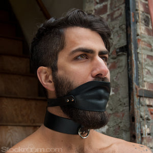 A dark-haired man’s face is shown in front of a stone wall. He has facial hair and is wearing the Over The Mouth Gag With A Silicone Ball and a black collar. The gag has a piece of black leather that completely covers his and a leather strap.