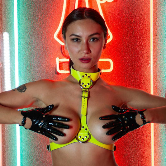 A nude woman with her hair pulled back wears the yellow Neon Angel Bust Harness and black latex biker gloves. She is standing in front of a glowing neon sign and covers her breasts with her hands.