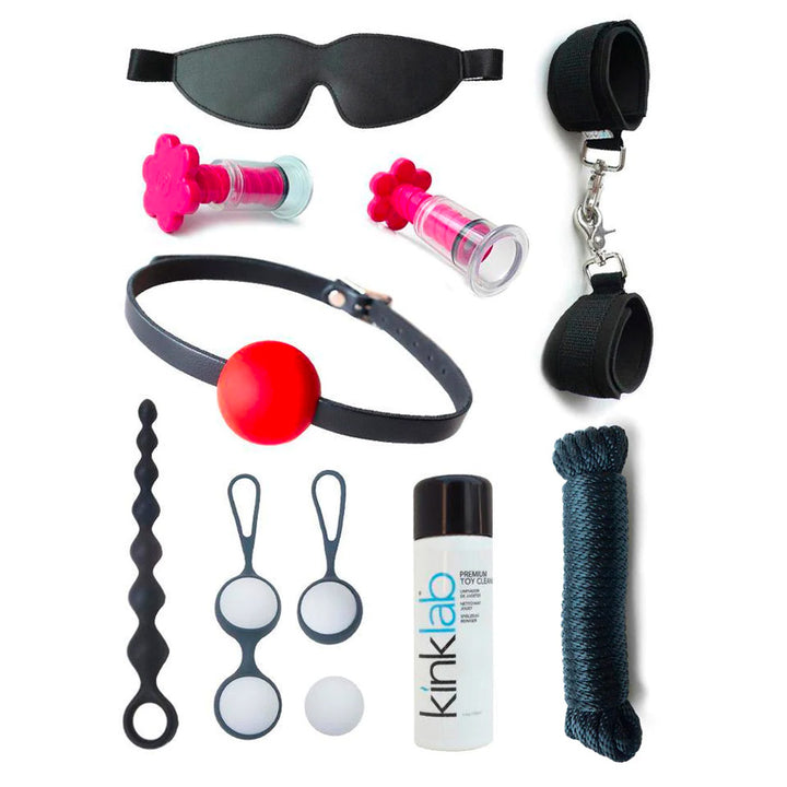 The contents of the Deluxe Stay At Home Adventure Kit are shown against a blank background. Shown is a black blindfold and wrist cuffs, black rope, pink suction T-cups, toy cleaner, a ball gag, anal beads, and kegel balls.