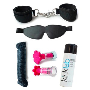 The contents of the Stay At Home Adventure Kit are shown against a blank background. Shown are black neoprene handcuffs, a black leather blindfold, black nylon rope, pink suction T-cups, and KinkLab toy cleaner.
