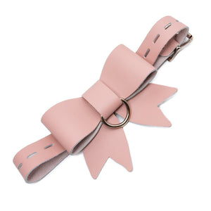 A product shot of the Stupid Cute Bow Restraint made by The Stockroom against a white background. The product is tilted diagonally against the background. It's made of light pink leather that comes together in a bow. There is a nickel-plated ring at the center of the bow. 