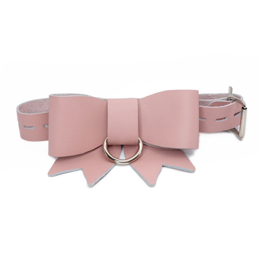 A product shot of the Stupid Cute Bow Restraint made by The Stockroom. It's made of light pink leather that comes together in a bow. There is a nickel-plated ring at the center of the bow. 