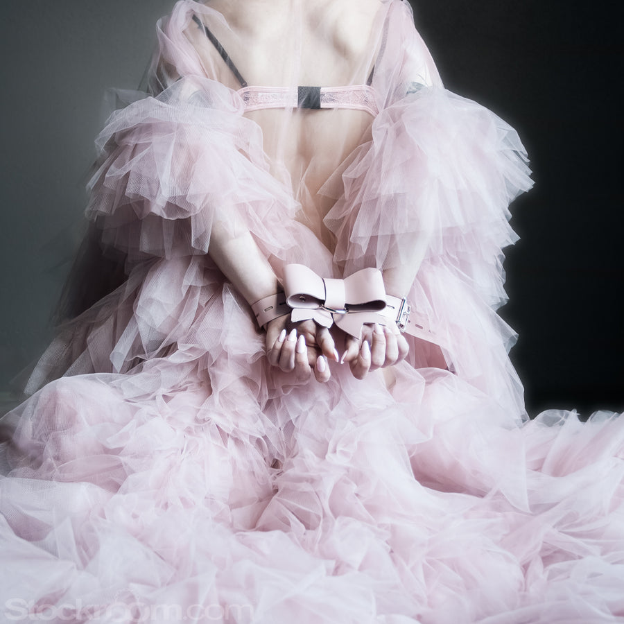 A model stands with their back toward the camera in a long, lacy dress that cascades onto the floor. Their hands are fastened behind their back with the Stupid Cute Bow Restraint, made of powder-pink leather with nickel-plated hardware. 