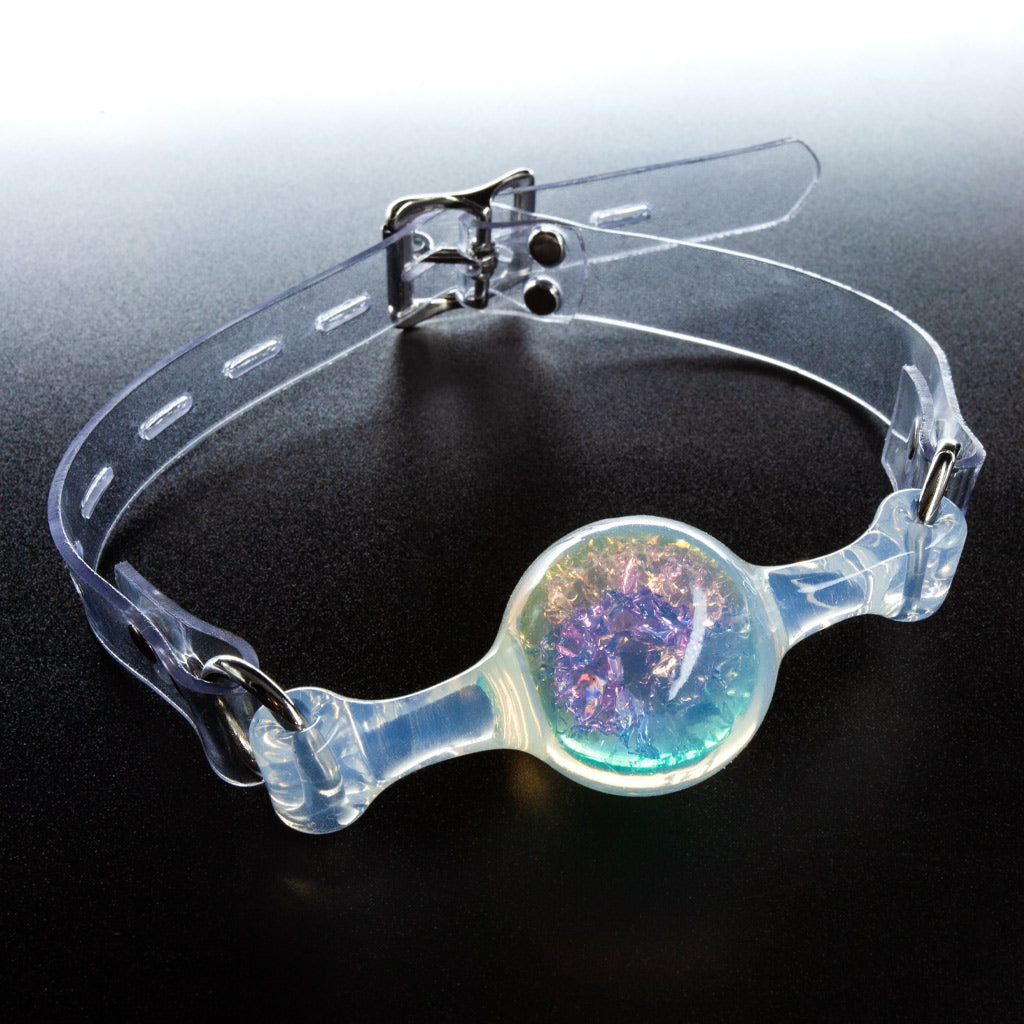 A ball gag made of transparent PVC rests on a black surface. The ball is filled with glittery, transluscent fragments of  purple, blue and yellow. 