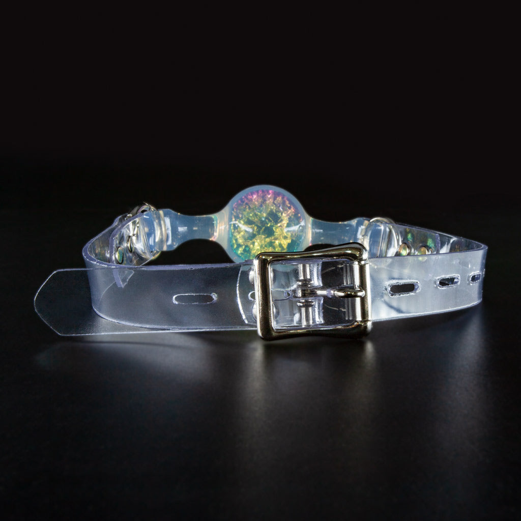A ball gag made of transparent PVC rests on a black surface. Viewed from behind, the focus is on the buckle and strap.