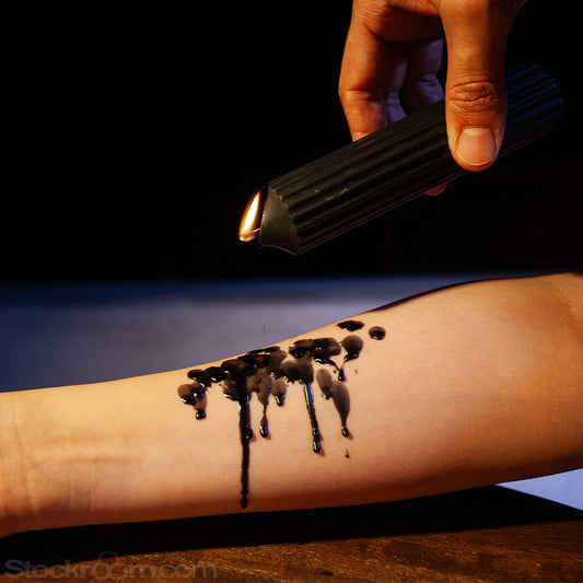 Close-up of a hand tilting a lit black scented wax play drip candle, dripping melted wax onto a person’s forearm, with glossy wax trails running down the skin against a dark background.