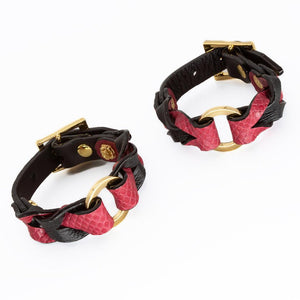 A pair of the Melanie Rose Designs x The Stockroom Wrist Cuffs, made of black and red leather braided together with a gold O-ring in the center, are displayed against a blank background.