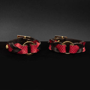 A pair of the Melanie Rose Designs x The Stockroom Wrist Cuffs, made of black and red leather braided together with a gold O-ring in the center, are displayed against a black background.