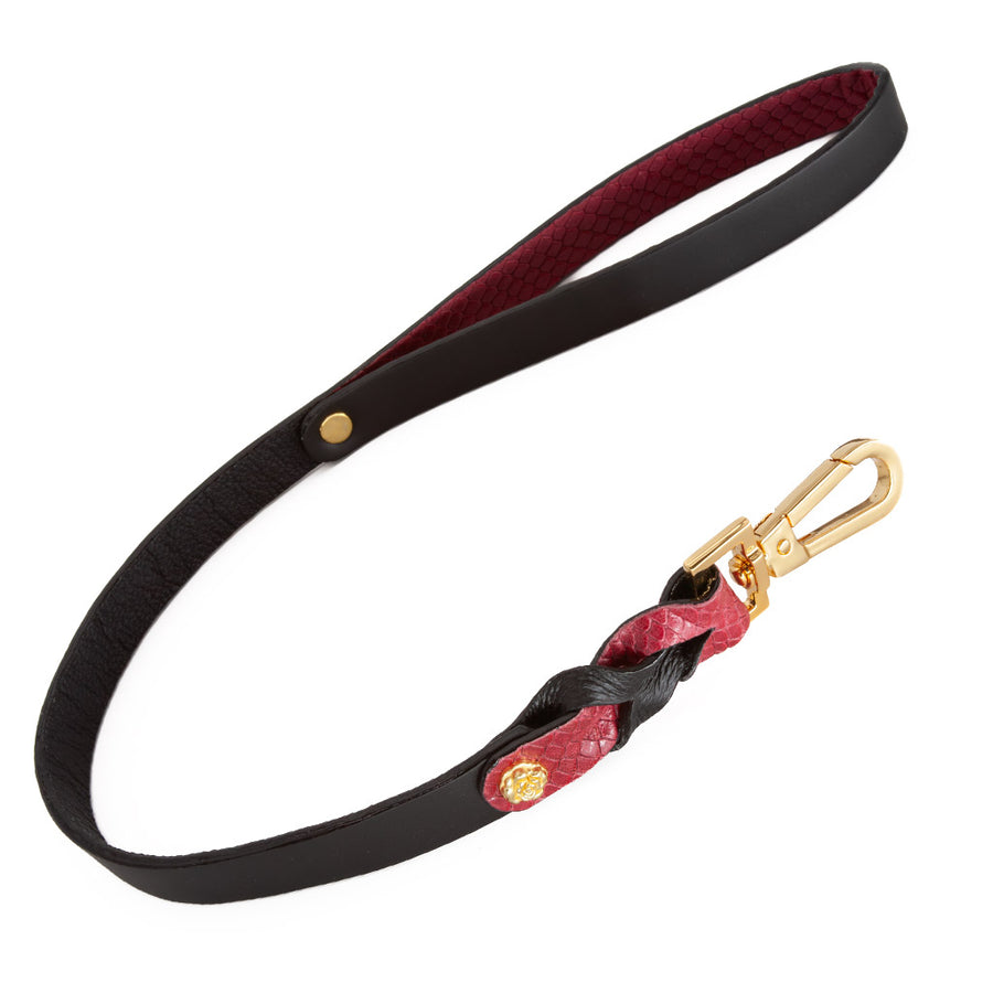 The Melanie Rose Designs x The Stockroom Leash is displayed against a black background. The leash is made of black leather with red braided leather accents at the bottom and a gold clip. The wrist loop is lined with red leather.