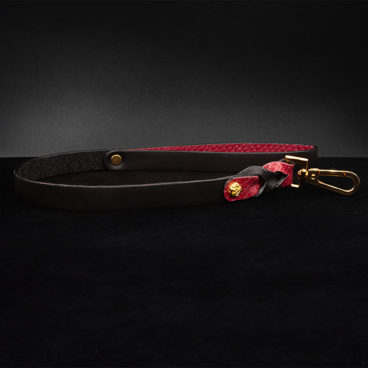 The Melanie Rose Designs x The Stockroom Leash is displayed against a black background. The leash is made of black leather with red braided leather accents at the bottom. The wrist loop is lined with red leather.
