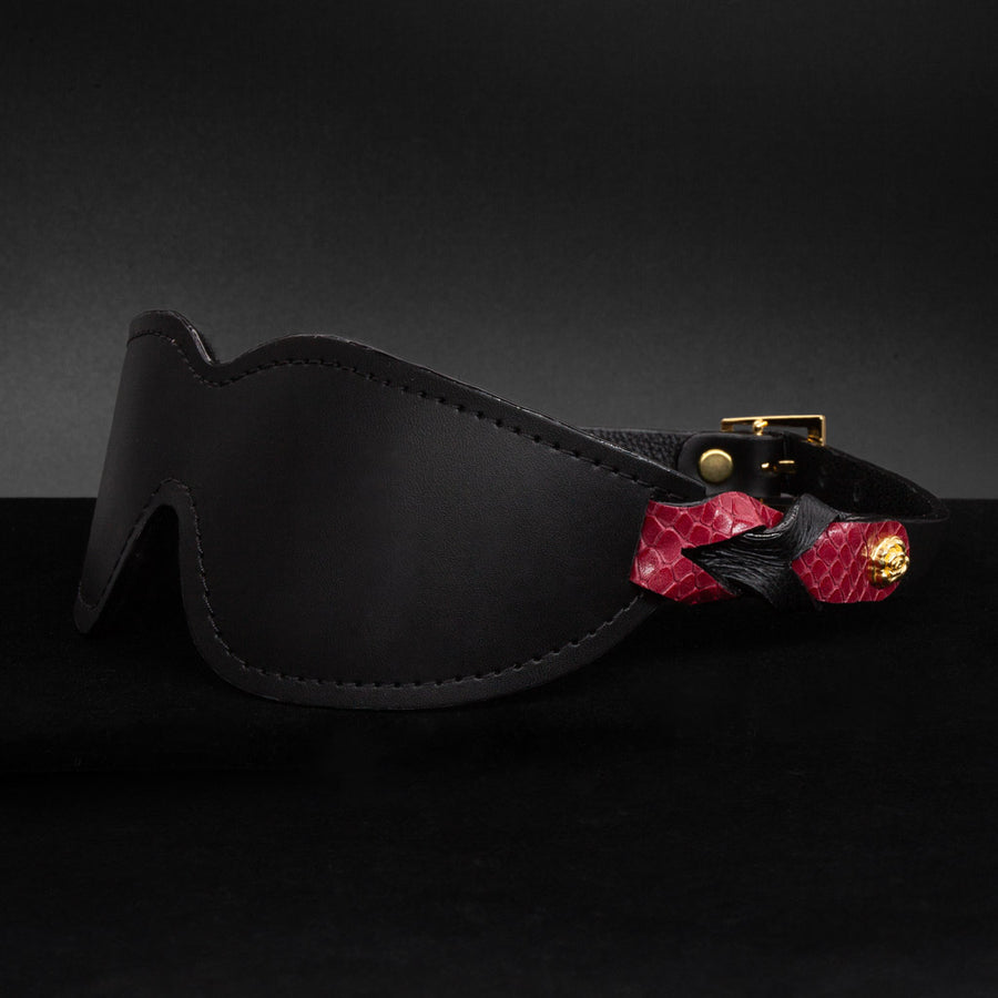 The Melanie Rose Designs x The Stockroom Blindfold, a black leather blindfold with straps made of braided black and red leather, is displayed against a black background.