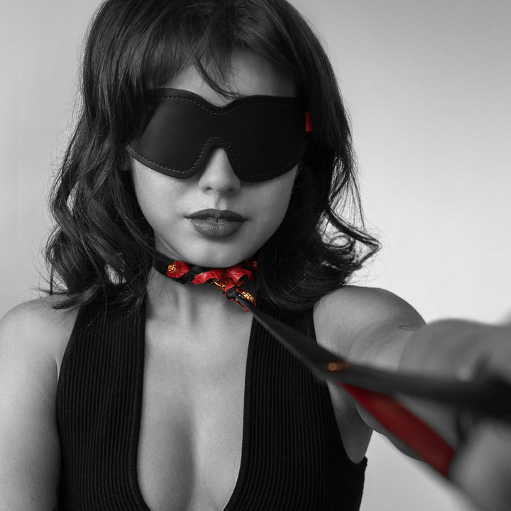 A woman with shoulder-length hair is shown wearing the Melanie Rose Designs x The Stockroom Blindfold. She wears a matching collar with a matching leash attached, which she holds out. 