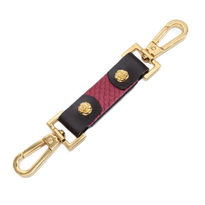 The Melanie Rose Designs x The Stockroom Restraint Clip is displayed against a blank background. It is made of a piece of textured red leather with black accents and a gold clip on each side. There is a small gold rose on each end of the clip.