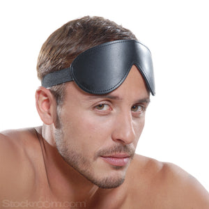 A close-up of a brunette man with light facial hair is shown against a blank background. He has the black Kinklab Bondage Basics Padded Leather Blindfold pulled above his eyes.