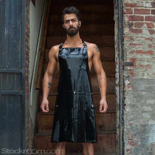 A muscular brunette man with facial hair stands in front of a wooden staircase. He wears the black Rubber Apron by Syren Latex, which has pockets and an adjustable neck strap. The hem of the apron ends at his knees.