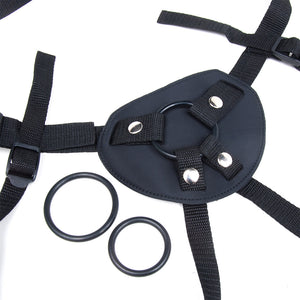 A close-up of the Malibu Terra Firma strap-on harness is shown against a blank background. One O-ring is attached to the harness, and two additional differently-sized O-rings are placed next to the harness.