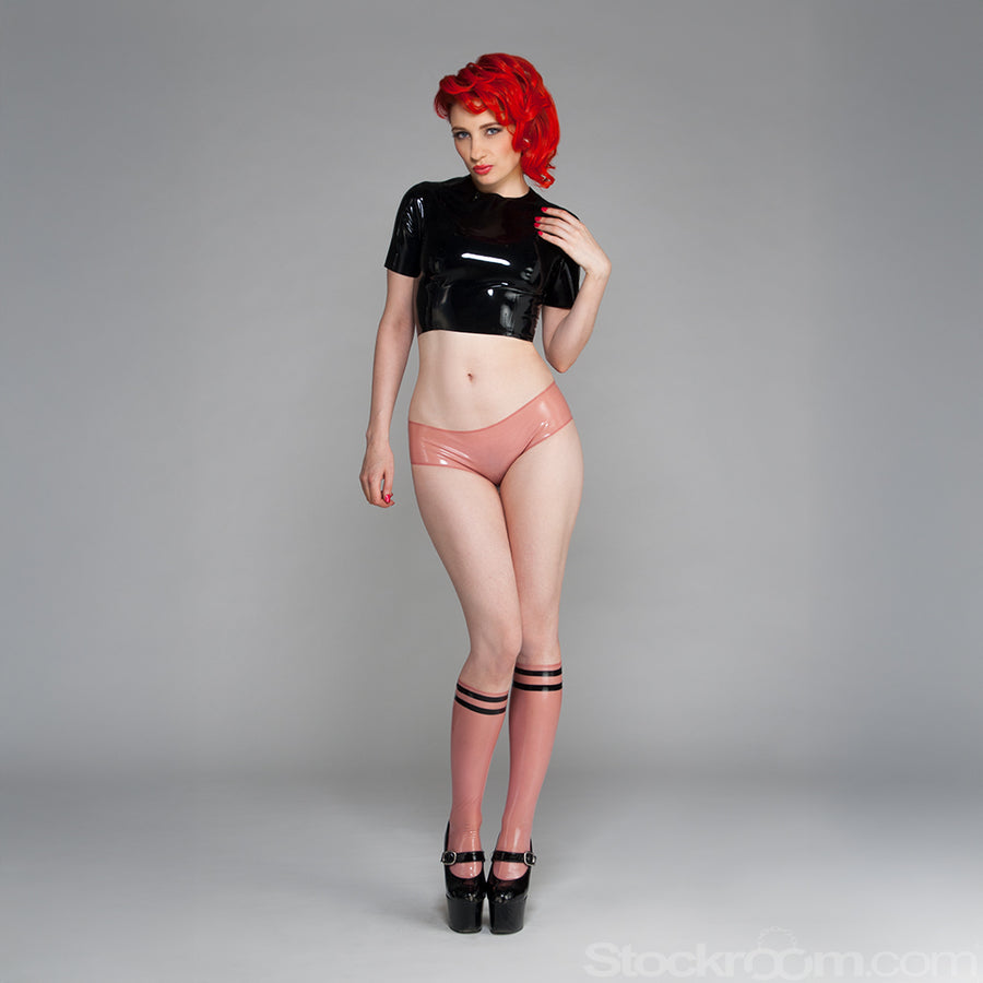 A woman with bright red hair poses in front of a grey backdrop. She wears a black latex crop top, pink latex panties, and the Varsity Socks by Syren Latex in transparent pink with black accents.