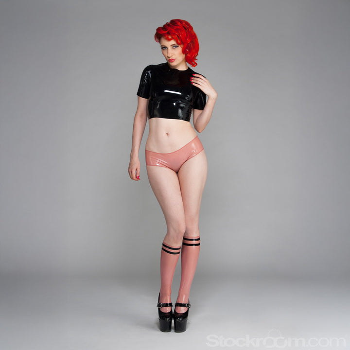 A woman with bright red hair poses in front of a grey backdrop wearing the Cheeky Panty by Syren Latex in rose pink. She also wears the latex zip-back crop top in black, athletic-style latex knee socks and black high heels.