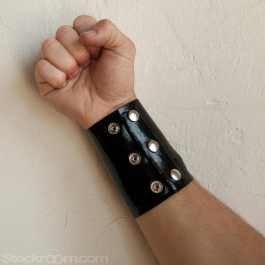 A close-up of a man's forearm is shown in front of a white wall. He wears the Latex Wrist Wallet by Syren Latex in black. The wallet resembles an armband and has two rows of three metal snaps.