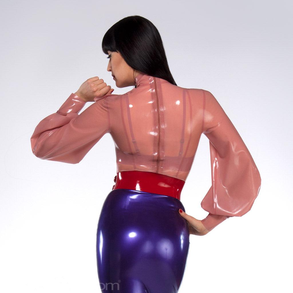 A woman with black hair poses in front of a grey backdrop, facing away from the camera. She wears the Garbo Blouse by Syren Latex in translucent pink, a purple latex pencil skirt, and a red latex waist cincher. The blouse has a zipper in the back.