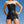 Load image into Gallery viewer, A woman stands with her hands on her hips with the blue sky behind her. She wears the black leather Corinthian Corset Dress, which is strapless and reaches the tops of her thighs. It laces up the front and has garter straps on the bottom edge.
