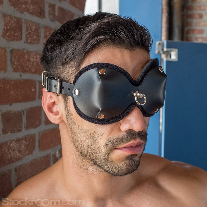  A close-up of a brunette man with light facial hair is shown. He wears the black Mistress Heather Leather Blindfold, which has cupped eye covers with a small metal D-ring between the eyes.